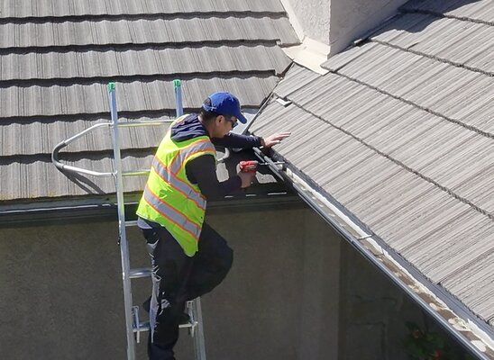 Ritchie on a ladder at a tile roof, fastening a gutter with a cordless drill
